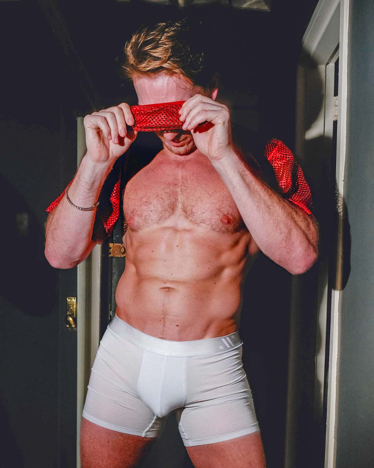 Man in white underwear holding a red object in front of his face, standing in a doorway.