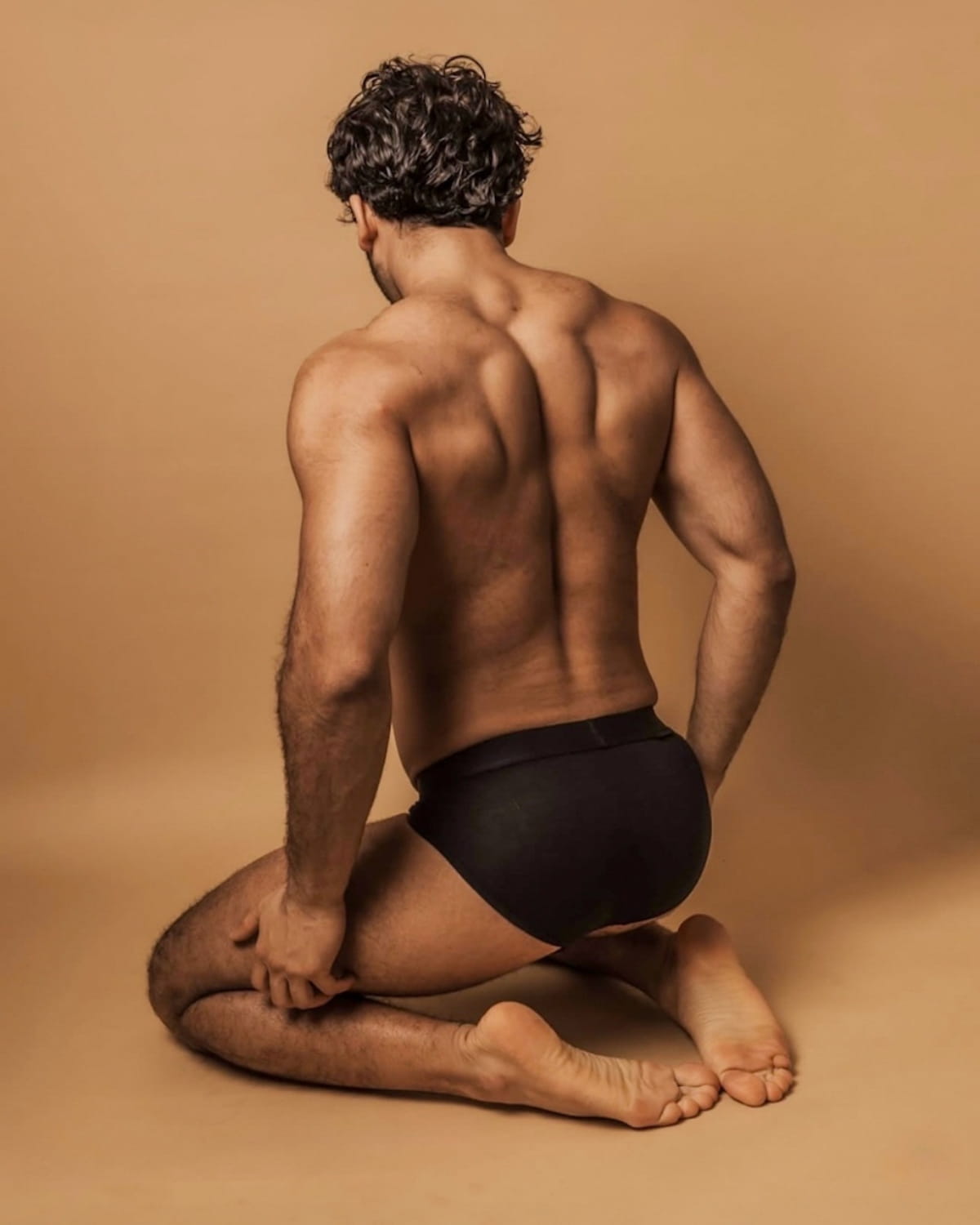 Man sitting with back to camera wearing black underwear on a beige background