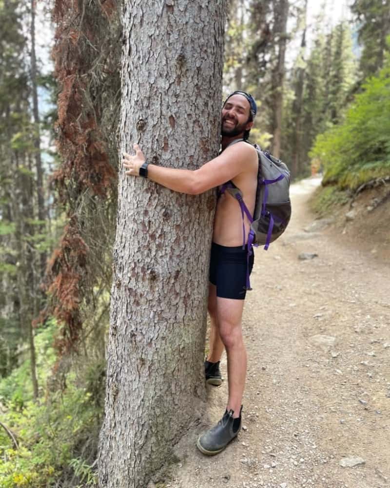 Smiling hiker in ALPHX underwear hugging a tree on a forest trail, symbolizing our sustainability journey and carbon offset impact through EcoCart.