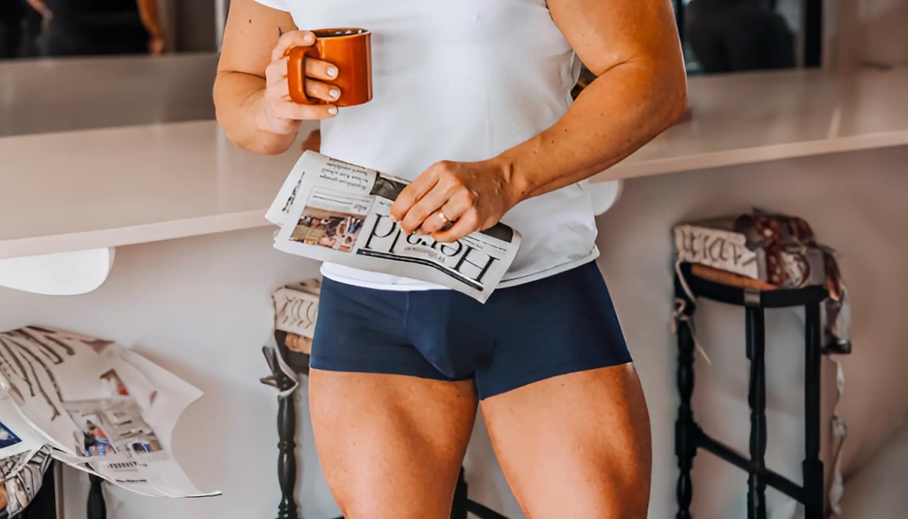 Person wearing a white t-shirt and navy shorts, holding a newspaper and a mug.
