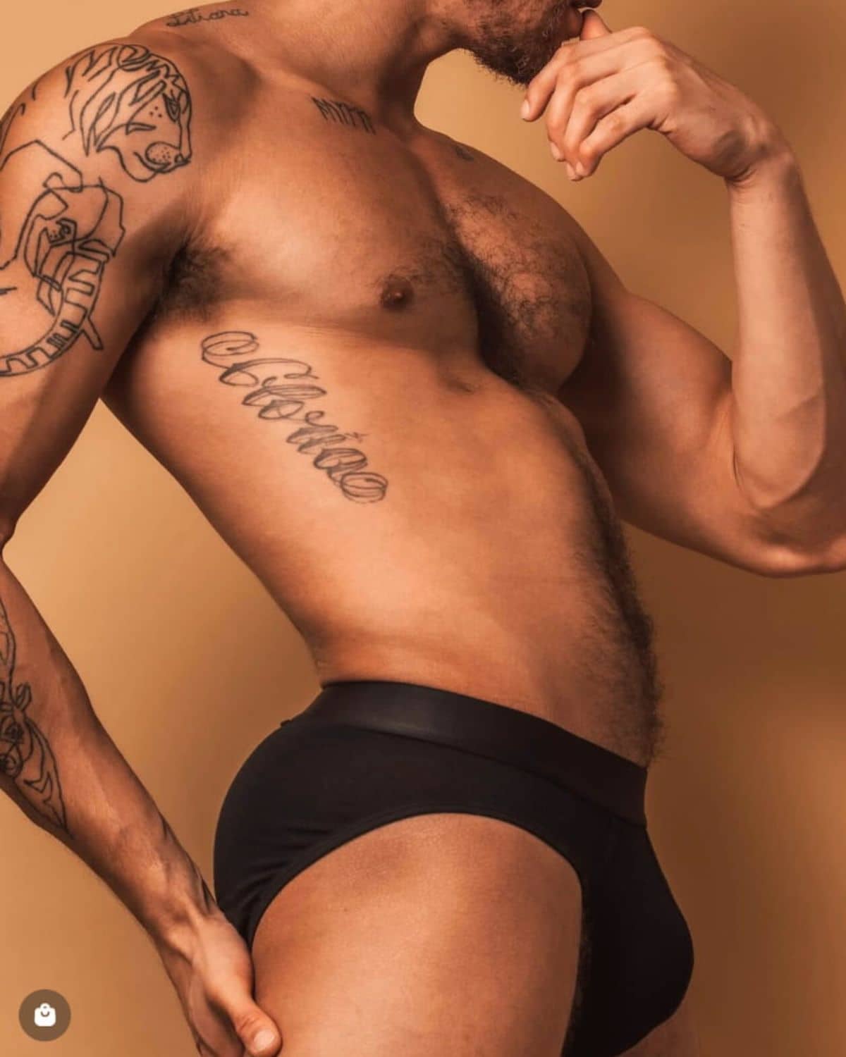 Male model wearing black ALPHX briefs poses against a warm tan background, highlighting the contoured fit, wide waistband, and sleek silhouette of the underwear.