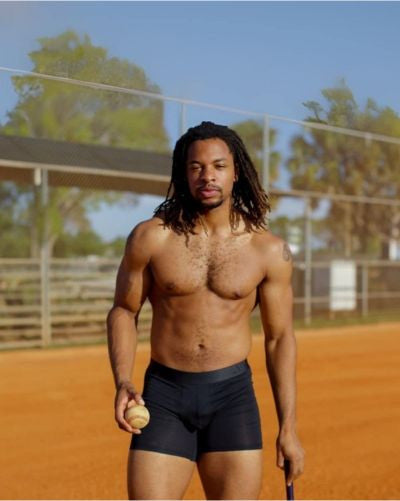 Man holding a baseball on a baseball field with trees and a building in the background