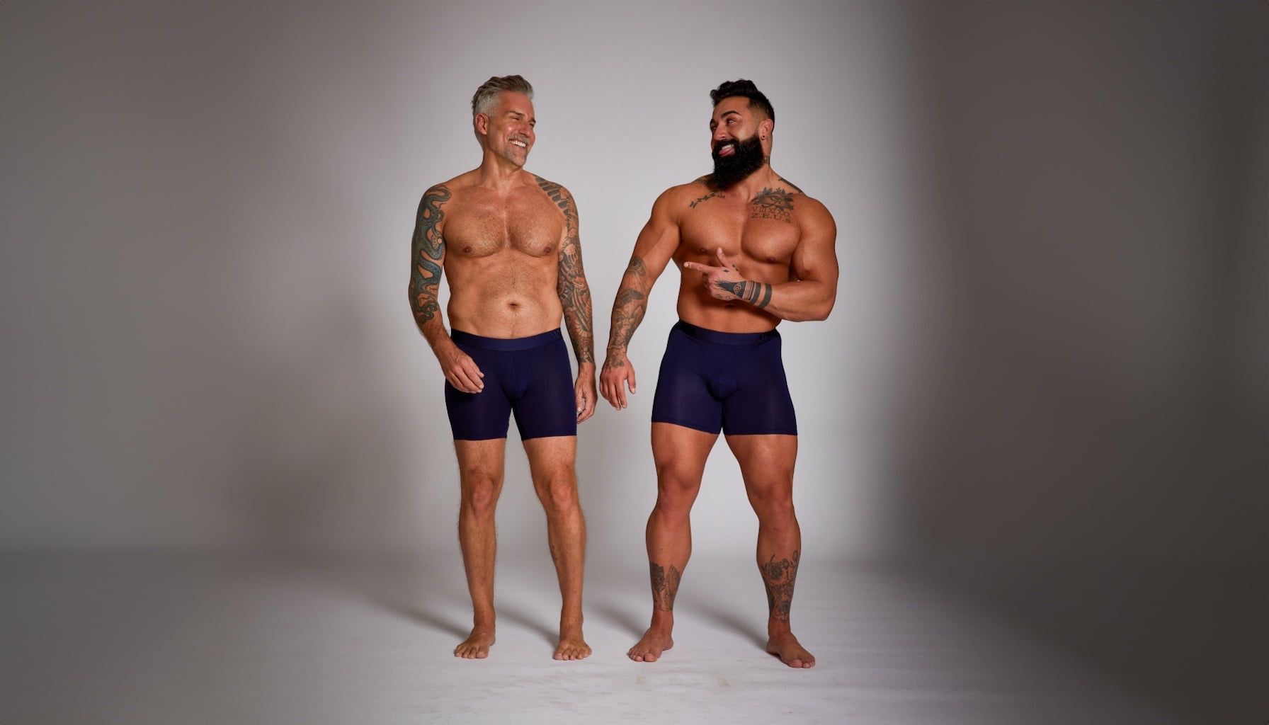 Two men wearing navy blue boxer briefs standing against a gray background