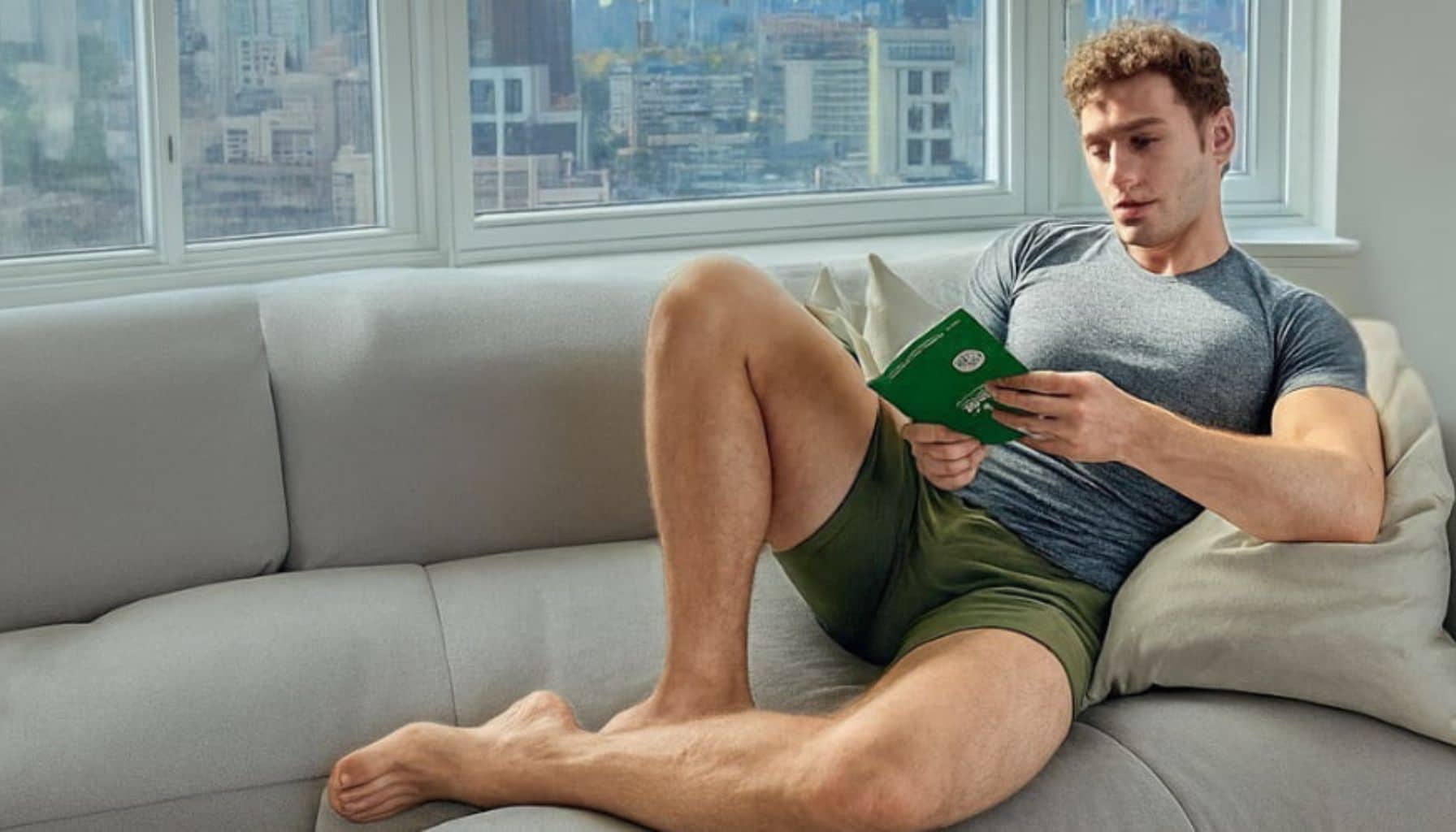 Man sitting on a couch reading a book with a cityscape wearing ALPHX Moss Green Boxer Briefs view through the window.