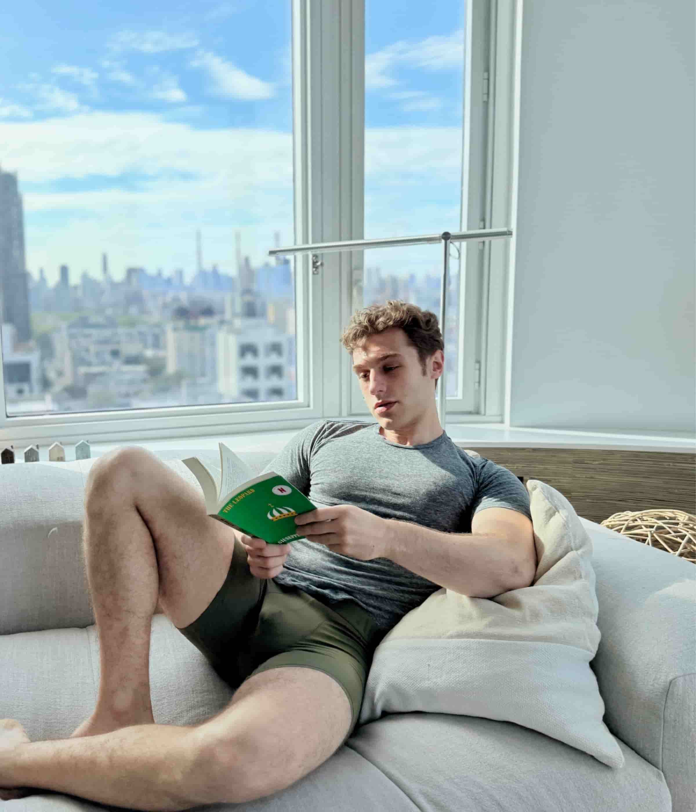Man reading a book on a couch with a cityscape view through the window